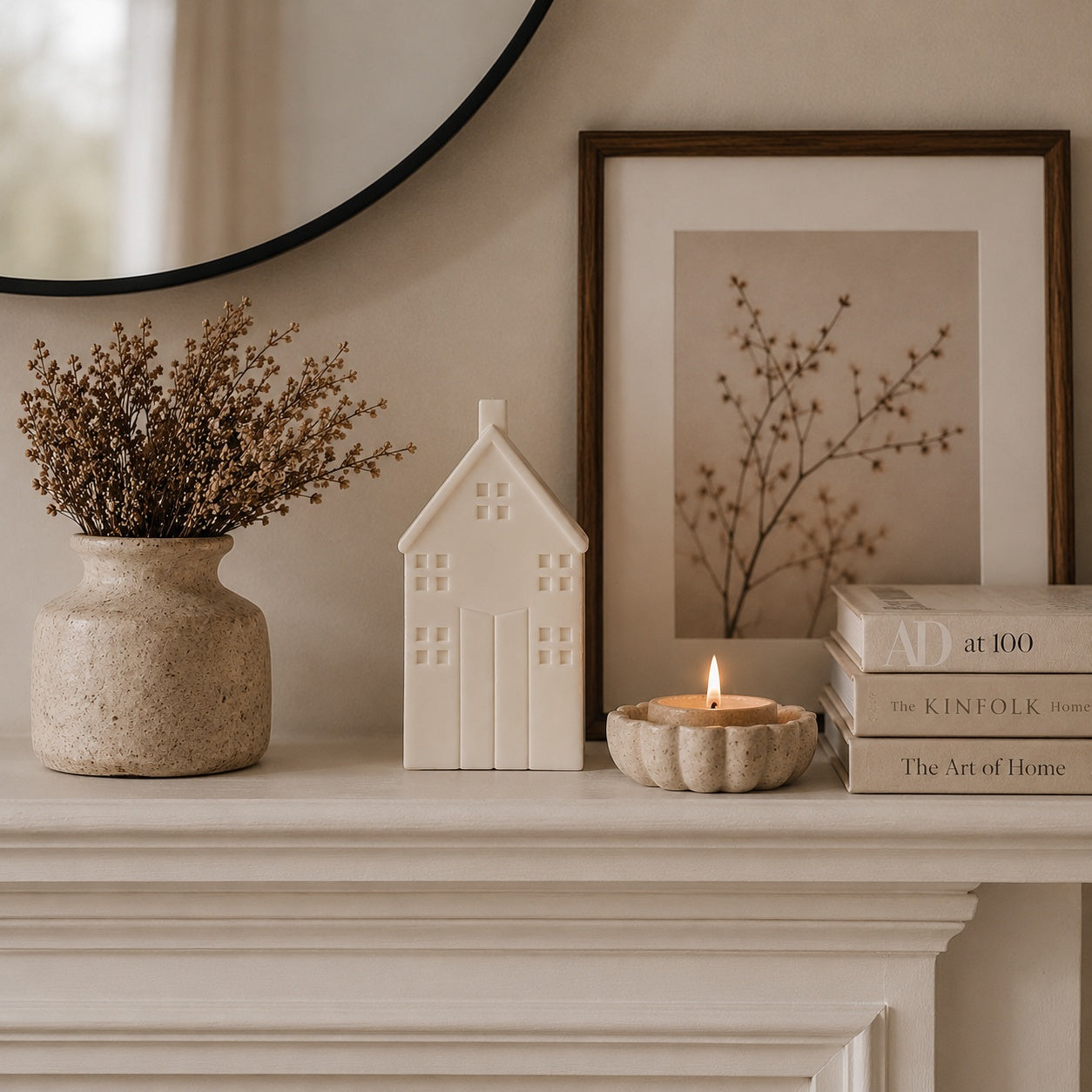 A styled interior scene on a mantel with a beige ceramic vase of dried branches, a small white house-shaped candle , a lit candle in a textured holder, and a stack of books, alongside framed wall art and a round mirror.