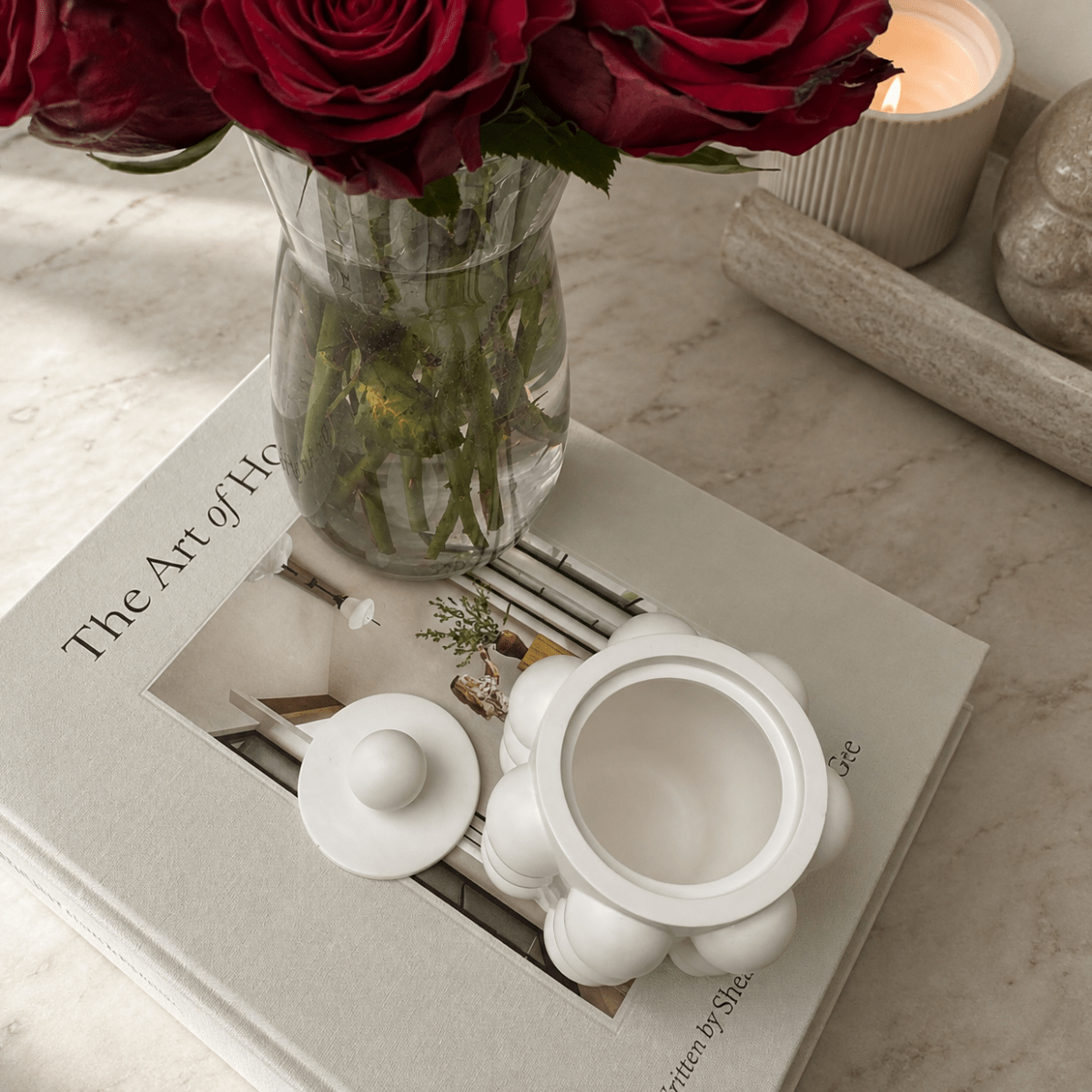 A white ceramic tureen with a matching lid sits on a book, styled with a glass vase of red roses and a lit candle on a marble surface.