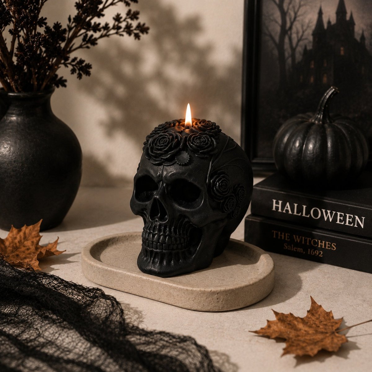 A black skull-shaped candle with rose details on top, lit and displayed on a stone tray in a Halloween-themed setting with books, a pumpkin, dried leaves, and black lace.