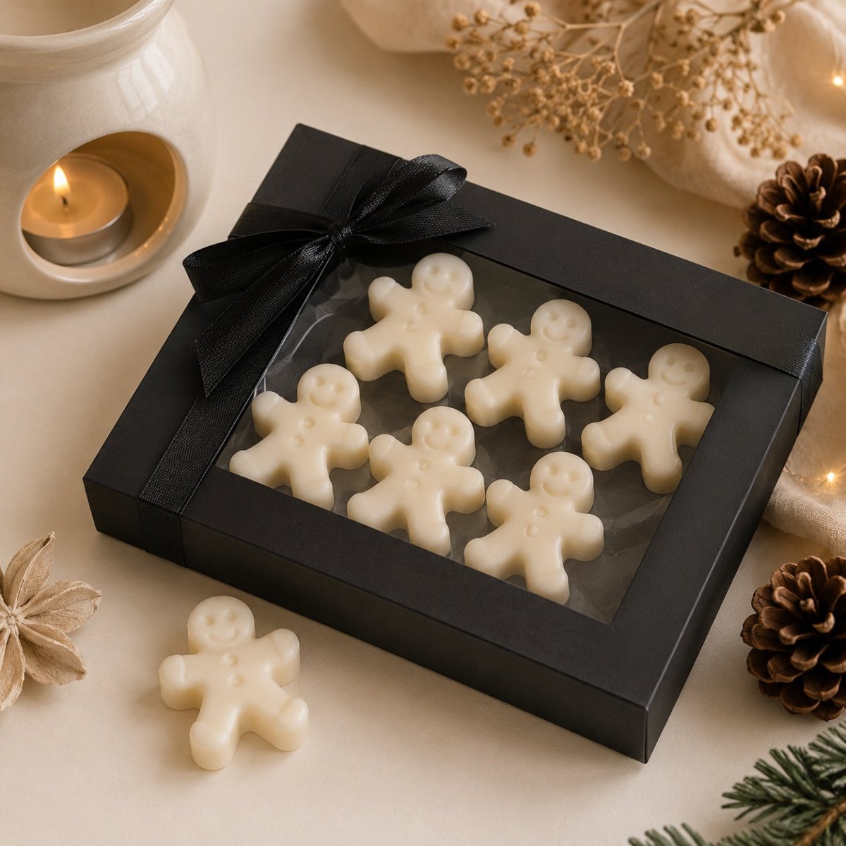 A black gift box with a clear window holding several cream-coloured gingerbread-shaped wax melts, tied with a black ribbon. A matching gingerbread piece sits outside the box on a warm neutral surface, with dried flowers, a candle, pinecones, and evergreen branches around it.