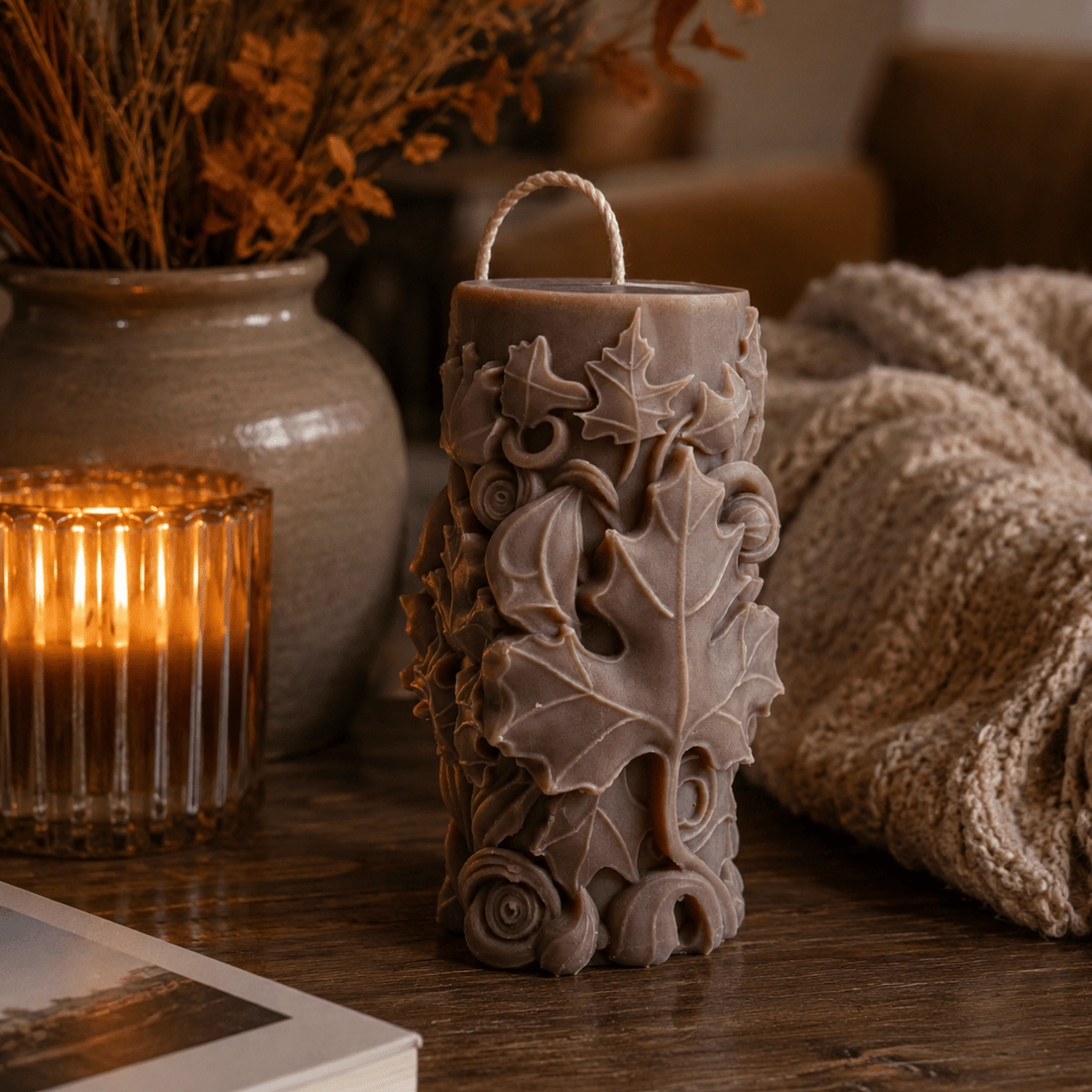 A carved pillar candle, featuring maple leaves, styled on a wooden table with a lit candle, a ceramic vase with dried flowers, and a knitted blanket in the background.