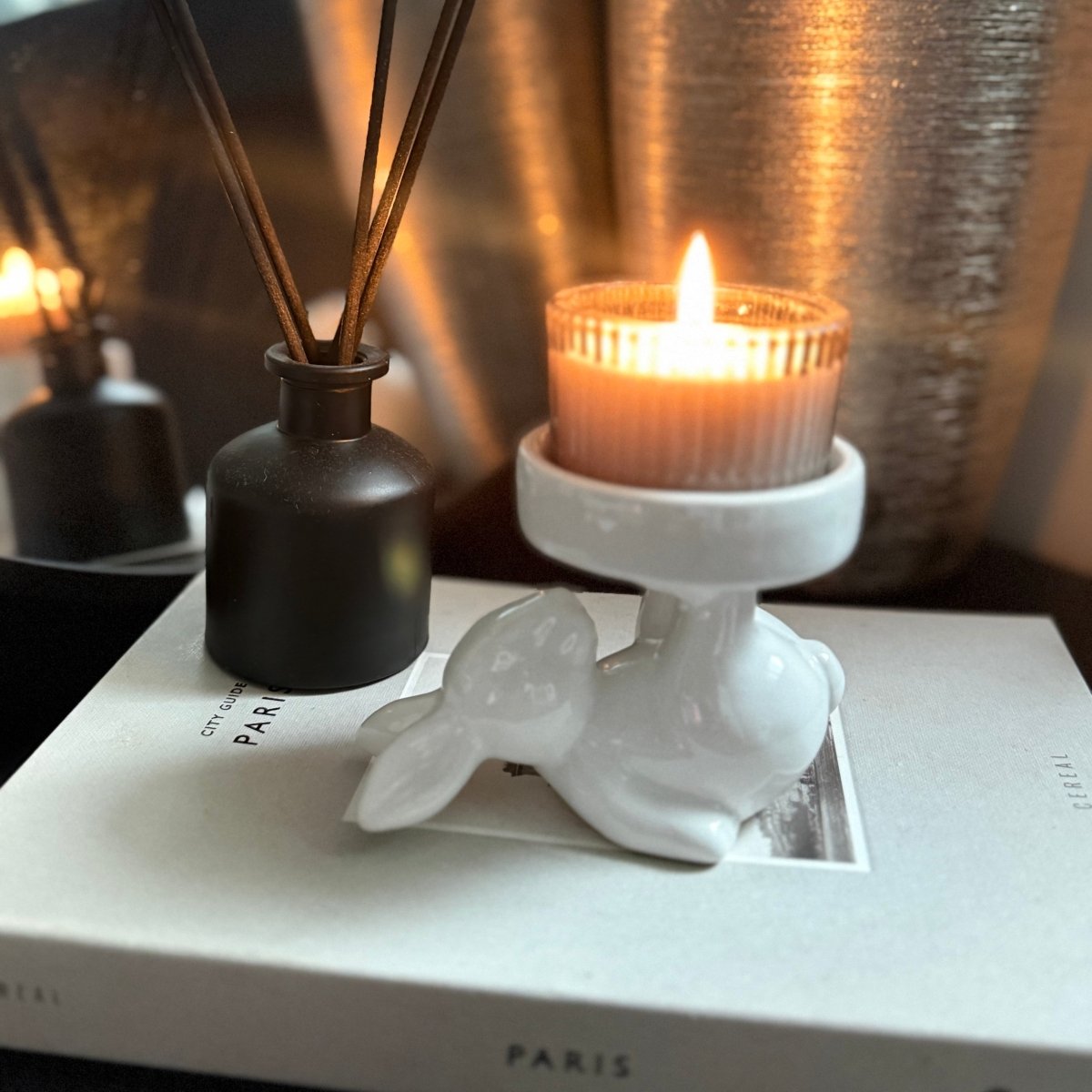white ceramic rabbit candle holder - Decorative setup with a candle, diffuser, and rabbit figurine on a book.