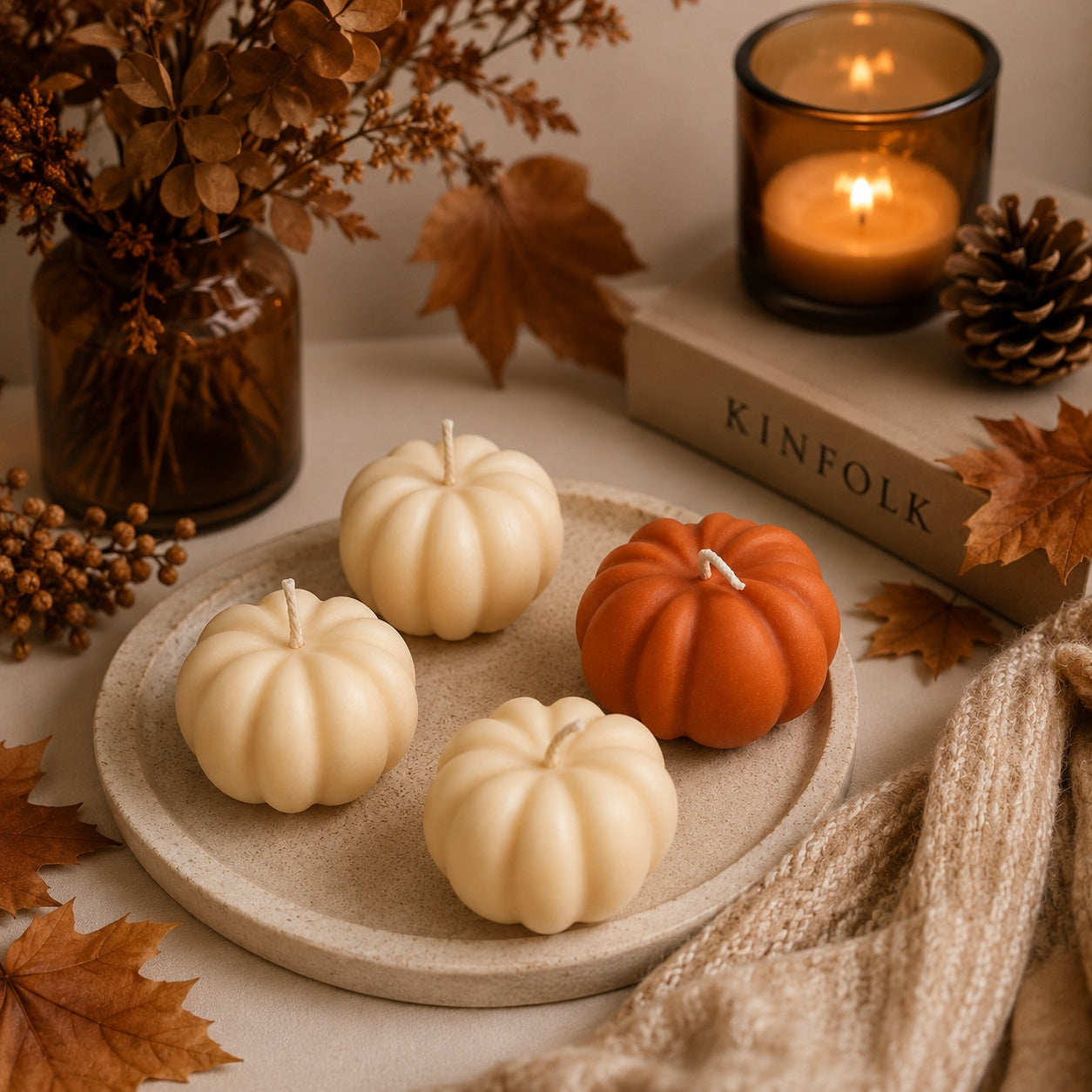 Four small autumn decorative pumpkin candles arranged on a round tray, including three cream-coloured pumpkins and one orange pumpkin, styled with autumn leaves, a candle, pinecones, and a book labeled KINFOLK.