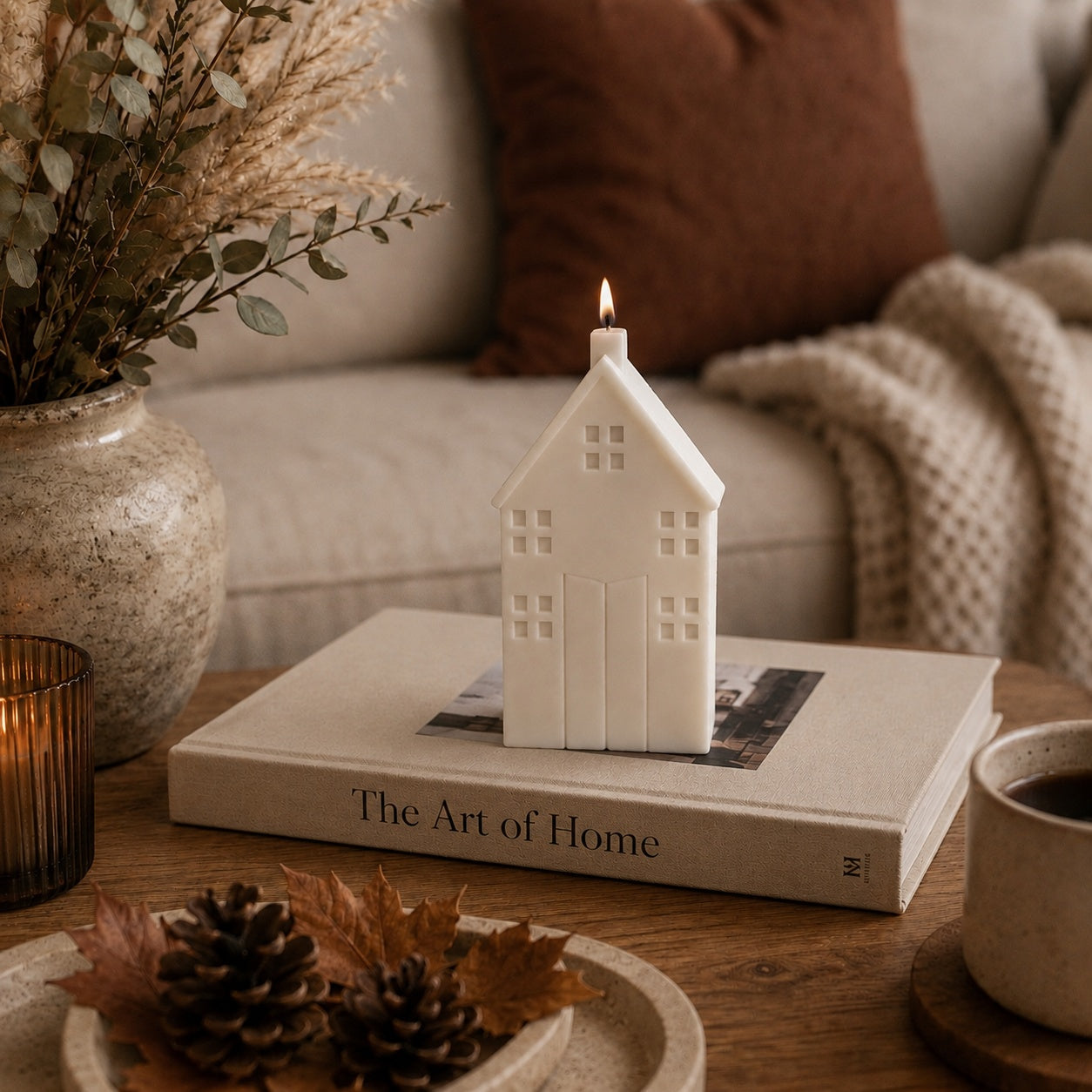 A white house-shaped candle with a lit flame sits on a book on a wooden table in a cosy autumnal living room setting.