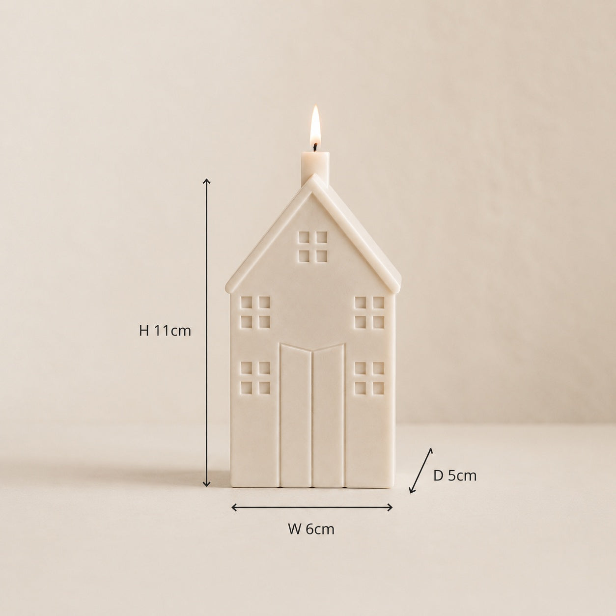 An off-white house-shaped candle with a lit flame, shown with dimension lines and labels indicating a height of 11cm, width of 6cm, and depth of 5cm against a neutral beige background.