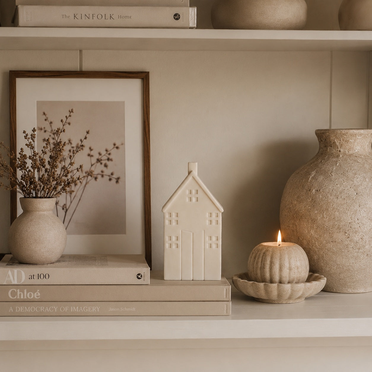 A styled shelf display featuring a small cream-coloured house-shaped candle, a matching flower-shaped saucer next to a textured ceramic vase with a lit candle, alongside books, a framed print, and a small vase with dried stems.