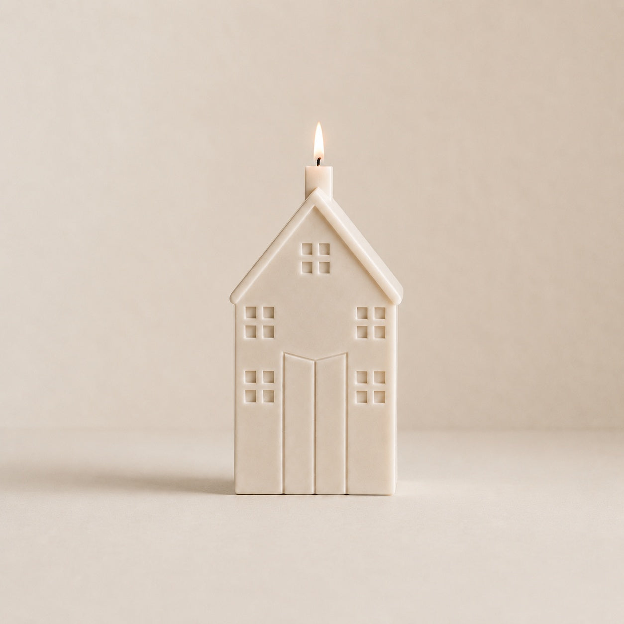 A house-shaped candle with a small flame burning from the top, made of a matte cream wax. The candle is centered against a solid, warm beige background.