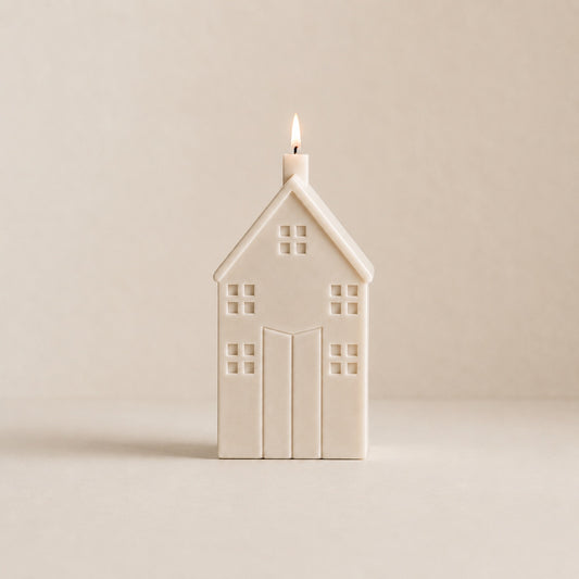 A house-shaped candle with a small flame burning from the top, made of a matte cream wax. The candle is centered against a solid, warm beige background.