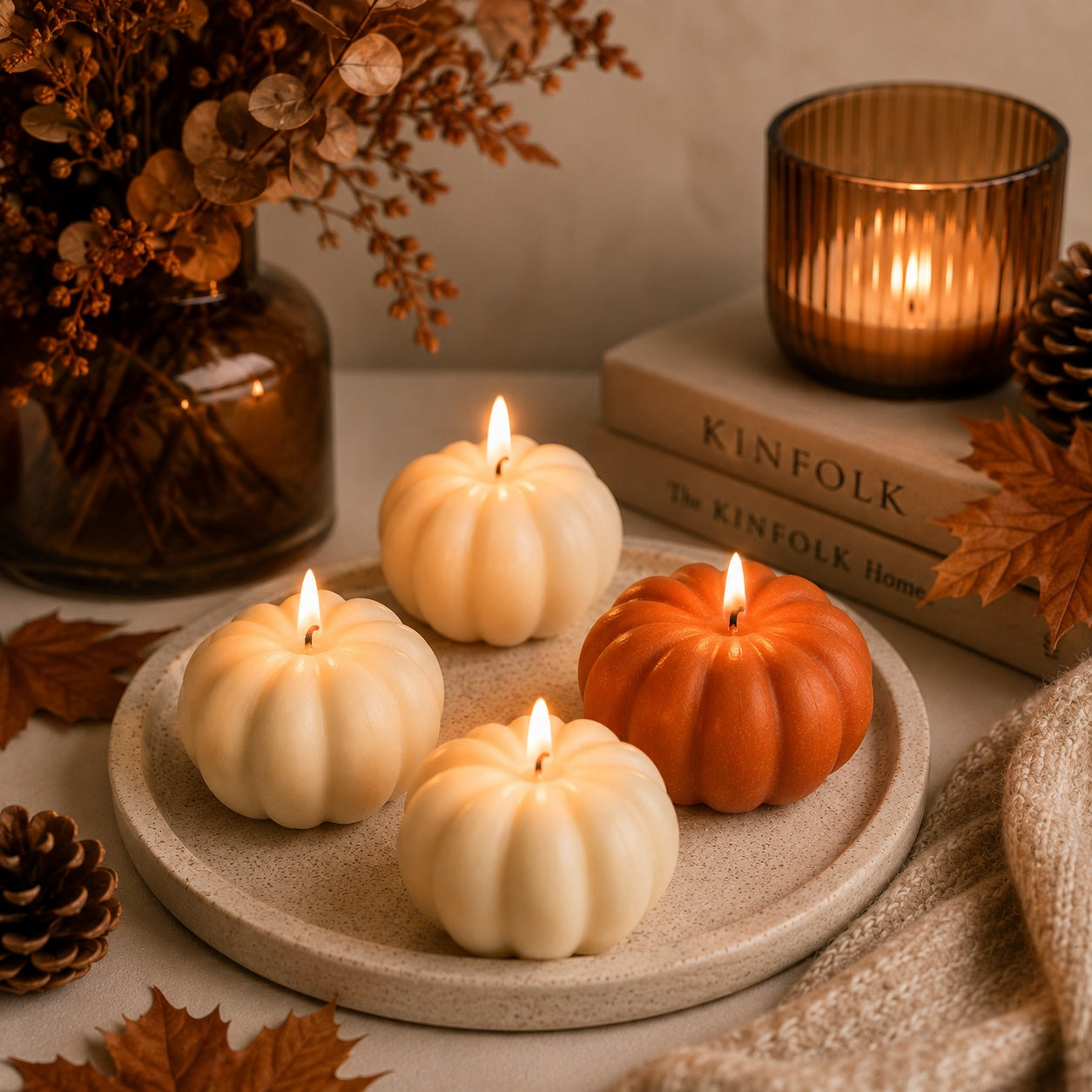 Autumn pumpkin hand-poured candles in cream and orange shades arranged on a round stone tray, styled with autumn leaves, pinecones, a lit amber candle in a glass holder, and a book in the background.