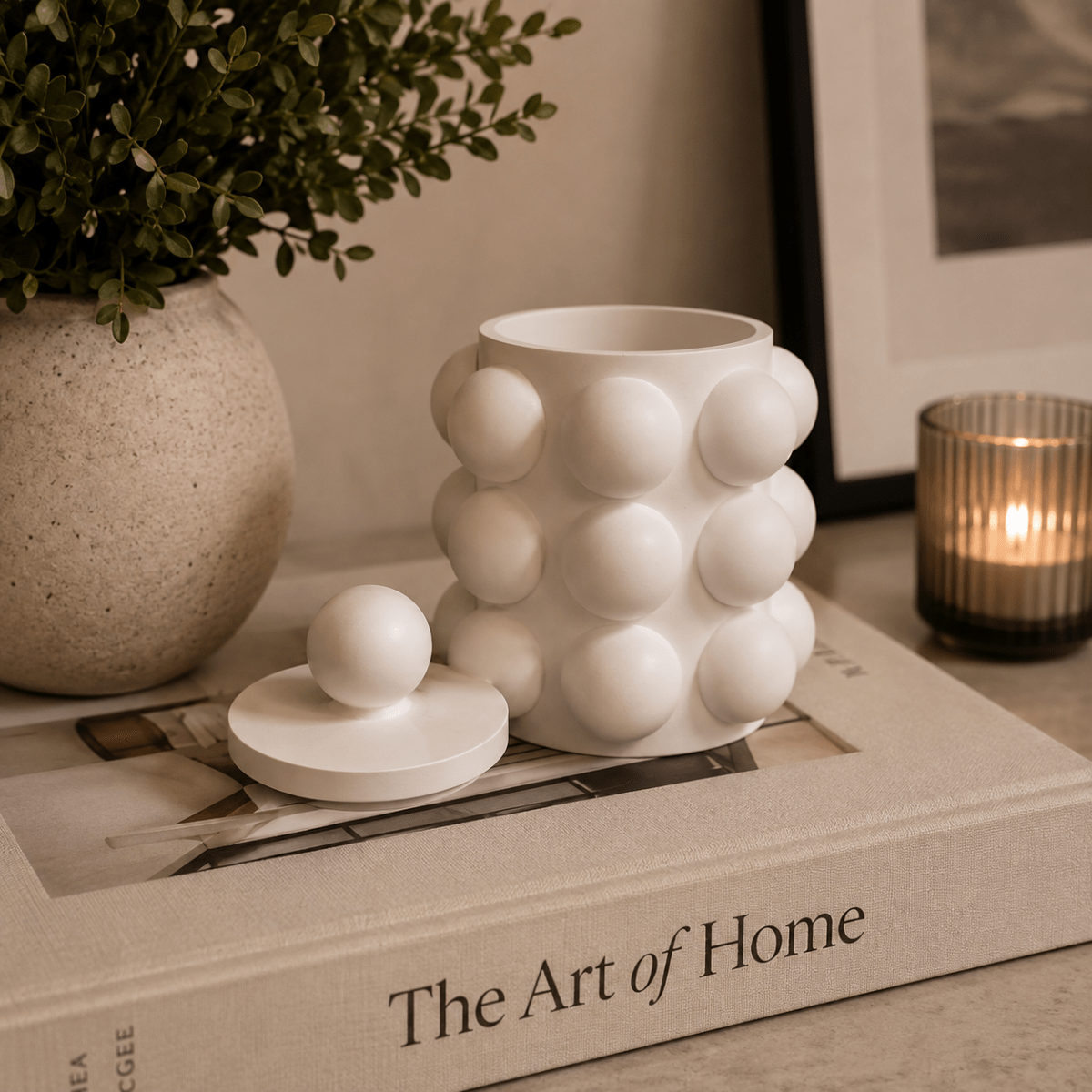 A white ceramic container with a matching lid, featuring a rounded, bubble-like textured pattern, displayed on a book in a styled home setting.