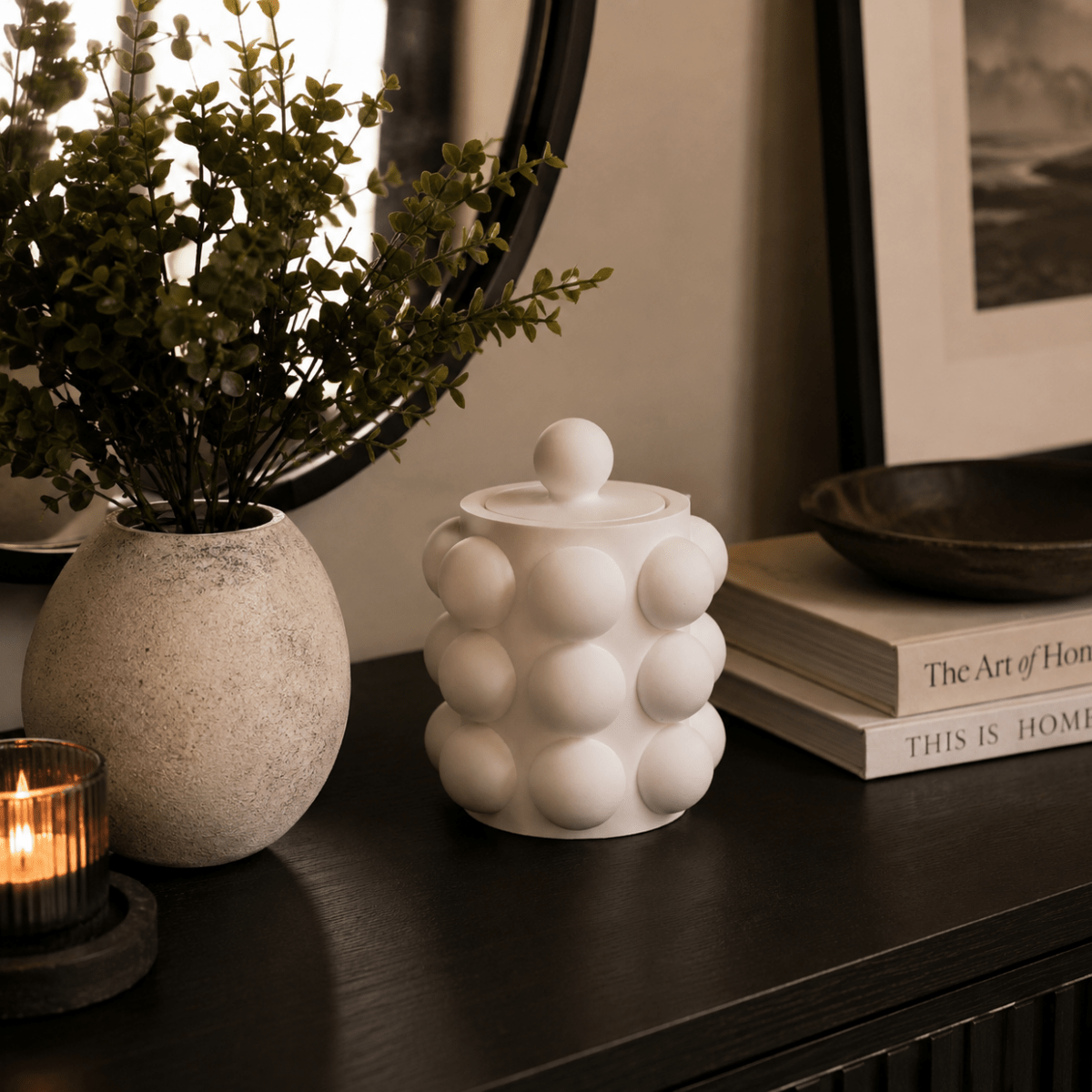 A white ceramic lidded container with a rounded bubble pattern sits on a dark wooden console table, styled with a textured vase of greenery, a lit candle, and books.