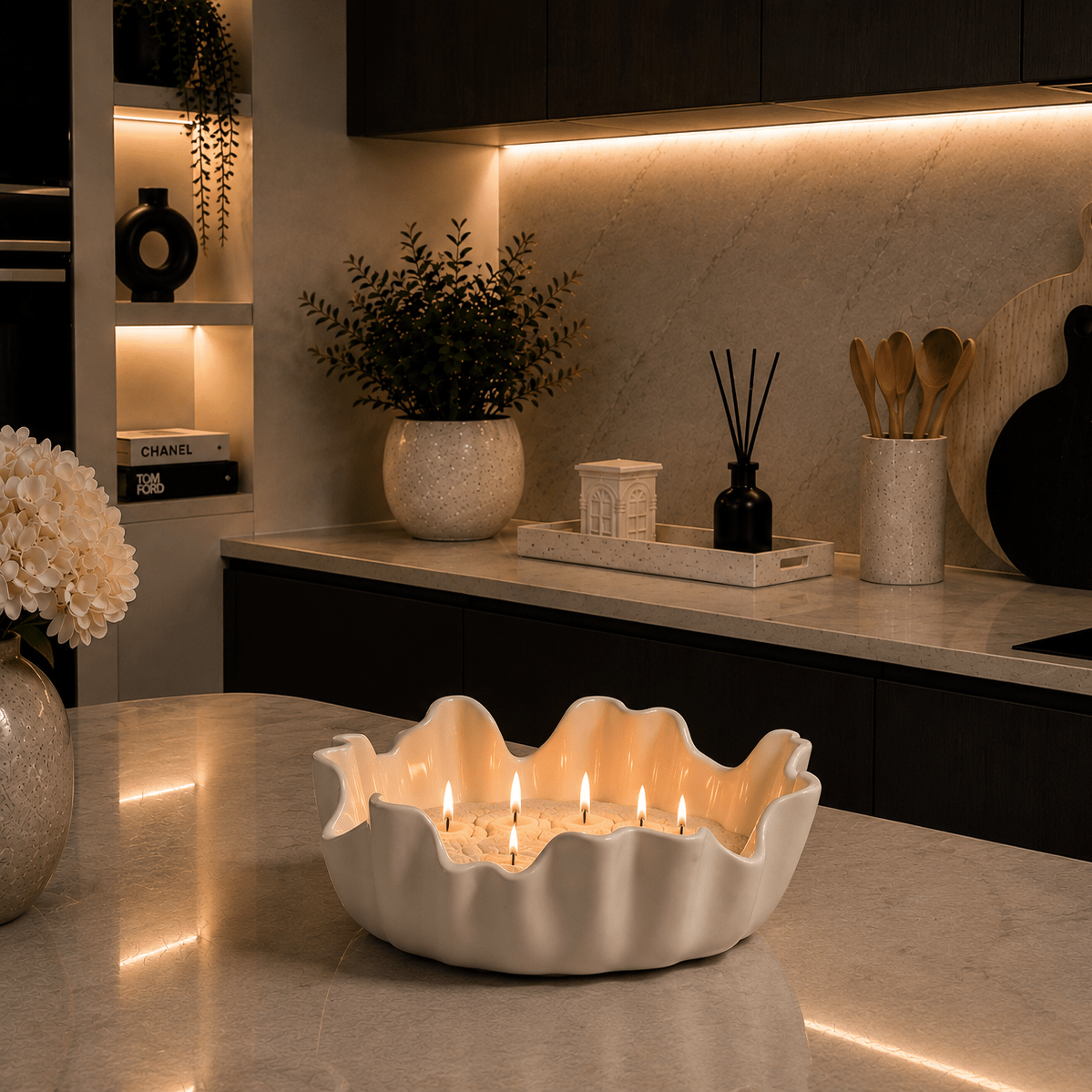 A white ceramic candle holder with a scalloped rim sits on a marble kitchen counter, lit with several candles inside. The background shows a modern kitchen with dark cabinets, warm under-cabinet lighting, and decorative items including a potted plant, cutting boards, and a diffuser.