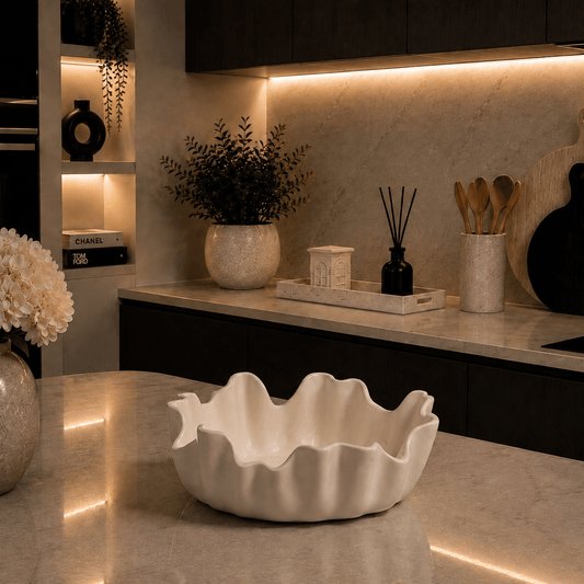 A white ceramic bowl with a wavy, scalloped rim sits on a polished marble kitchen island. The background shows a modern kitchen with dark wood cabinets, warm under-cabinet lighting, and decorative plants and utensils.