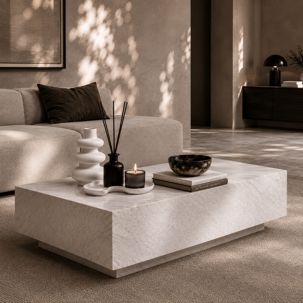 A modern low rectangular coffee table in a warm beige stone or concrete finish, styled in a minimalist living room with a cream sofa, candles, a reed diffuser, and a bowl.