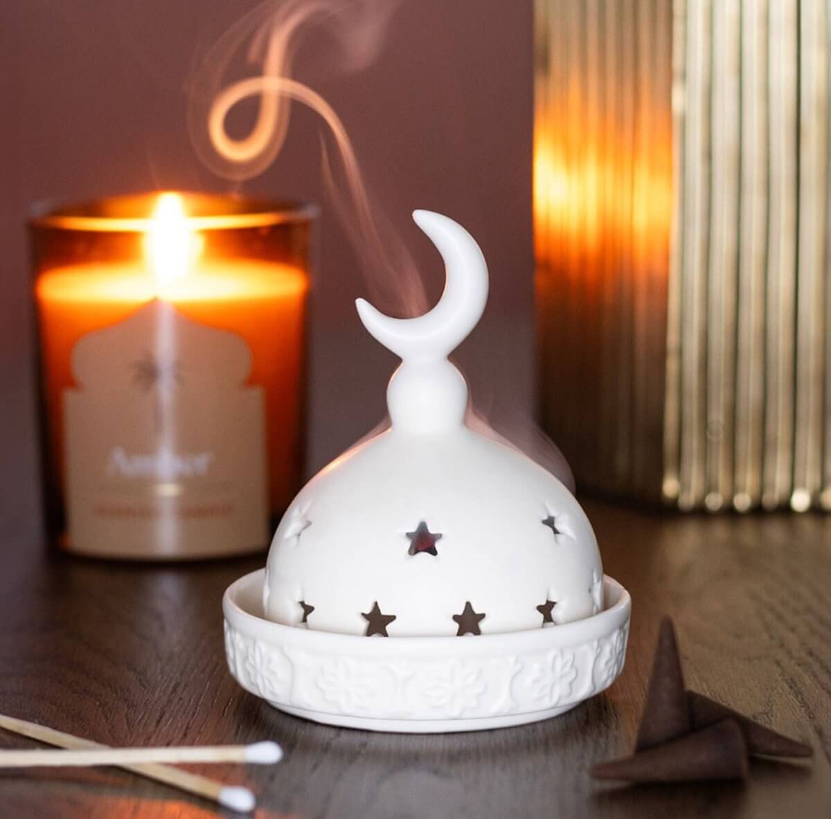 White ceramic incense burner with moon and star design on a wooden surface with a lit candle in the background.