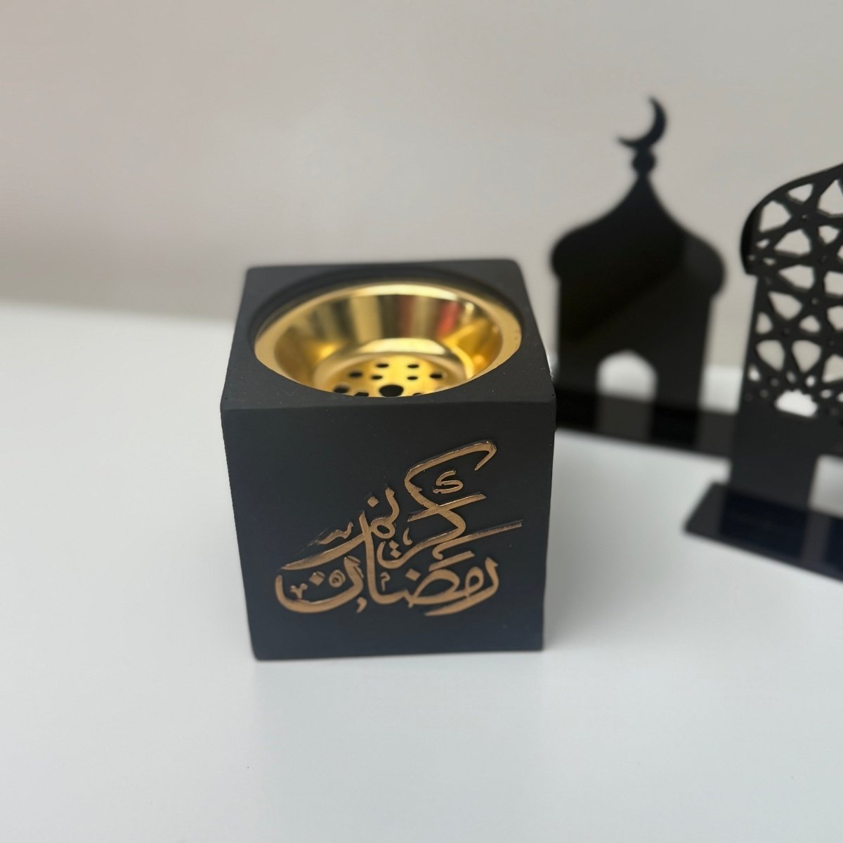 Black cube-shaped bakhoor incense burner  with gold interior and Arabic text on a white background