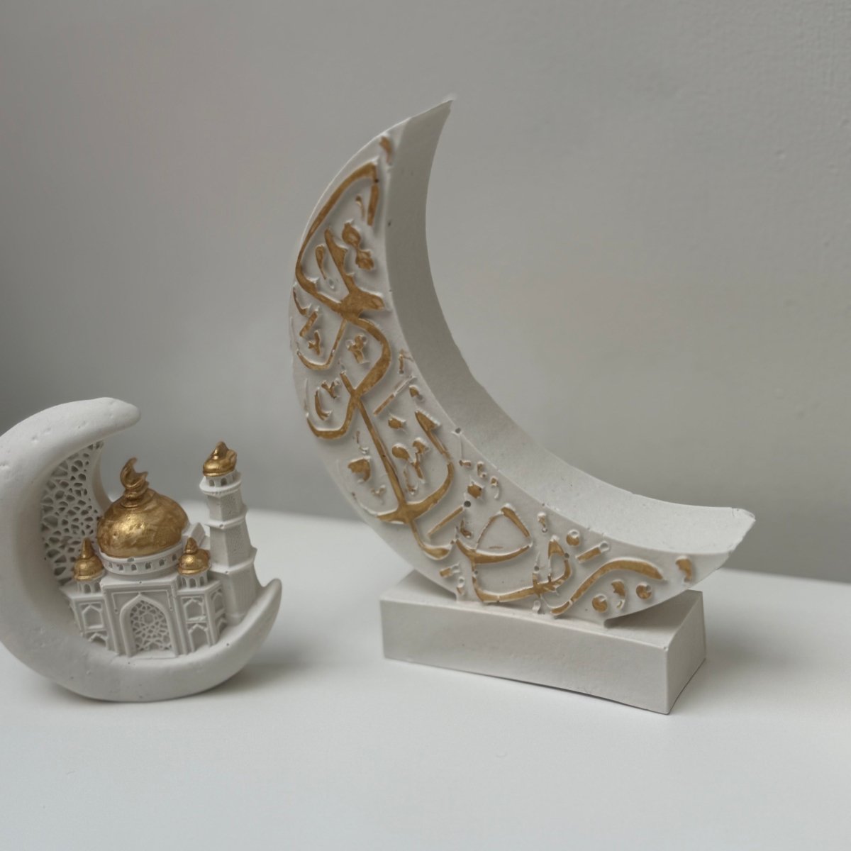 Decorative crescent moon with gold patterns and a small model of a mosque on a white surface.