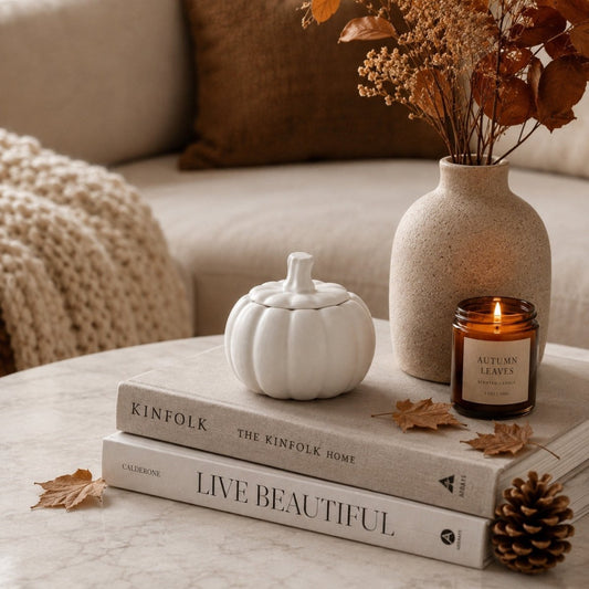 A styled autumn interior scene with a lit amber candle labeled 'Autumn Leaves' on stacked books, beside a white pumpkin-shaped container, a beige ceramic vase with dried leaves, and a pinecone on a light stone surface.