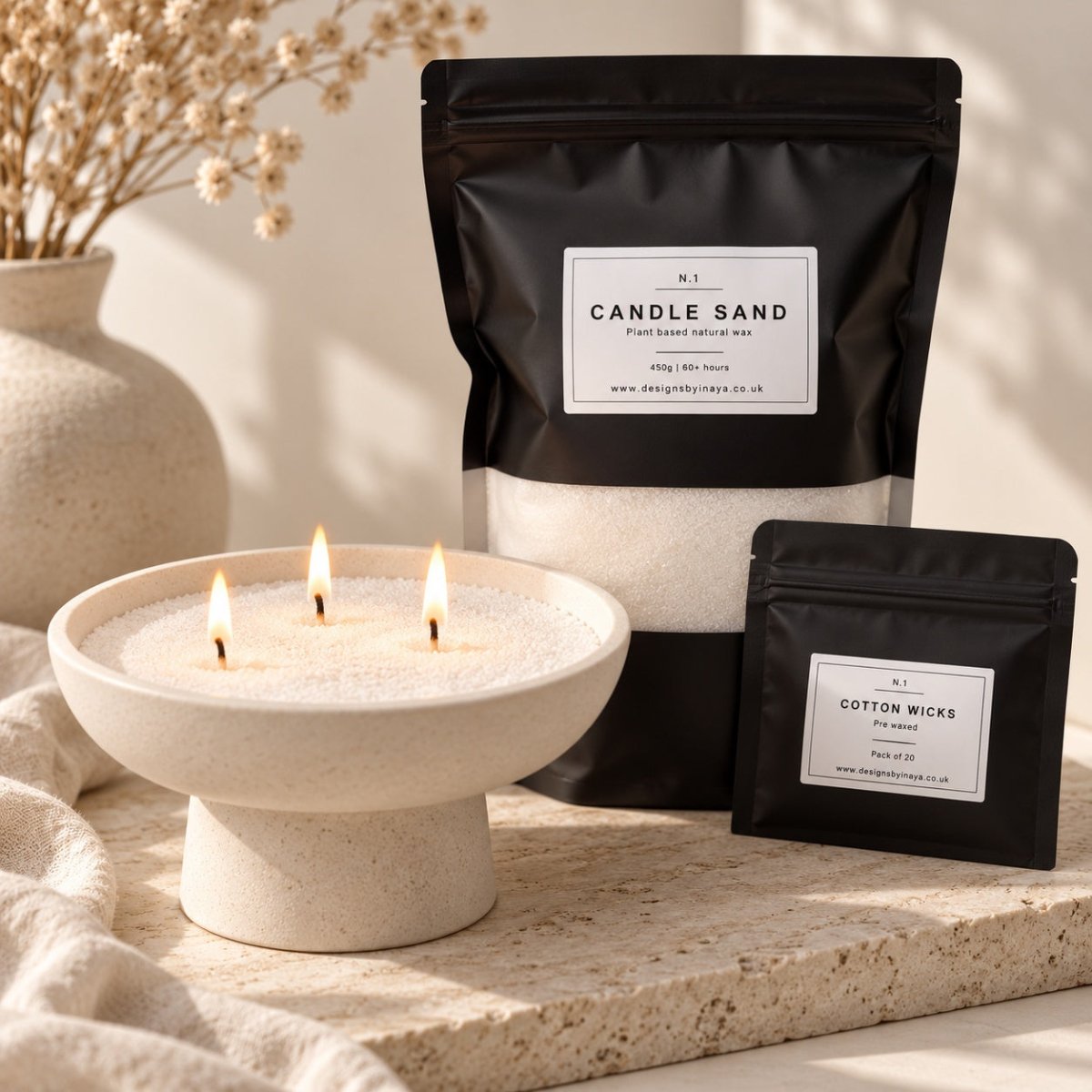 A styled product shot showing a three-wick candle in a cream pedestal bowl with lit flames, alongside two black resealable pouches labeled 'Candle Sand' and 'Cotton Wicks' on a stone surface with soft neutral props in the background.