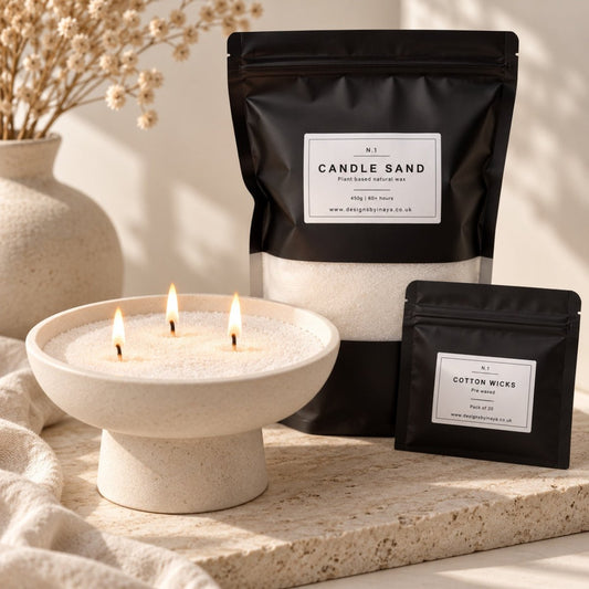 A styled product shot showing a three-wick candle in a cream pedestal bowl with lit flames, alongside two black resealable pouches labeled 'Candle Sand' and 'Cotton Wicks' on a stone surface with soft neutral props in the background.