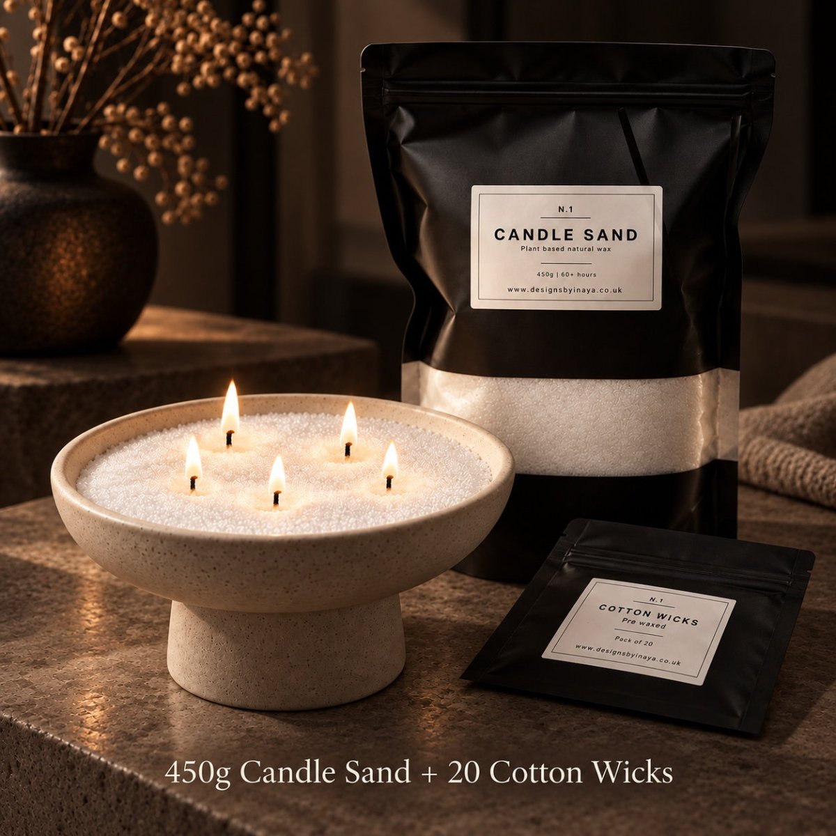 A styled product shot showing a black stand-up pouch labeled 'CANDLE SAND' behind a lit candle in a stone pedestal bowl, with a smaller pouch labeled 'COTTON WICKS' in front. The scene is set on a textured stone surface with warm ambient lighting and decorative dried stems in the background.