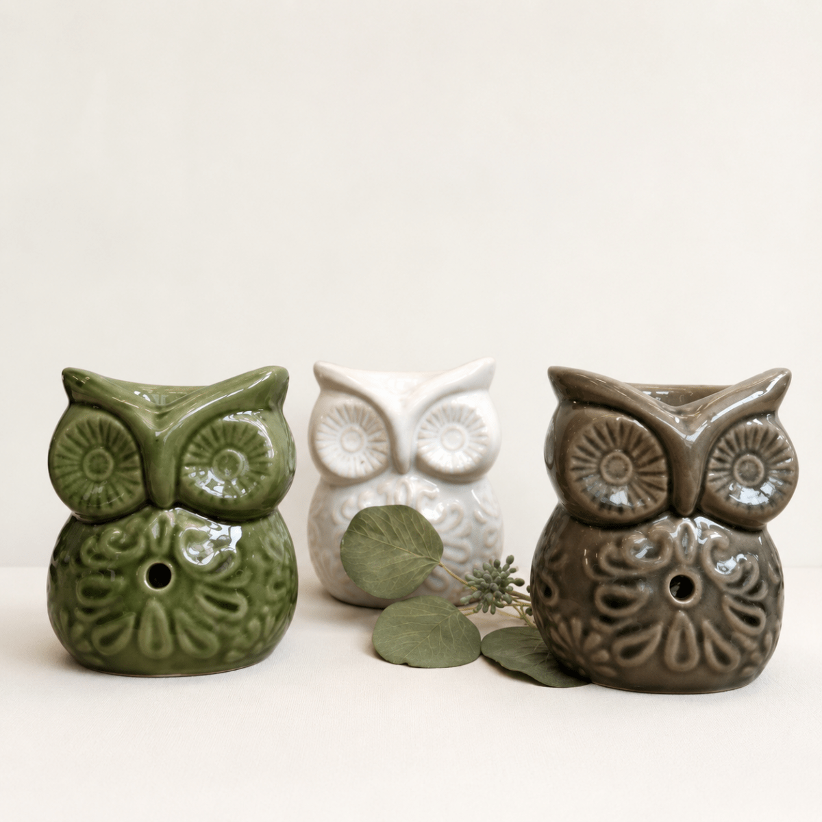 Three ceramic owl oil burners in green, cream and taupe displayed on a clean light neutral background with eucalyptus leaves. Autumn home styling