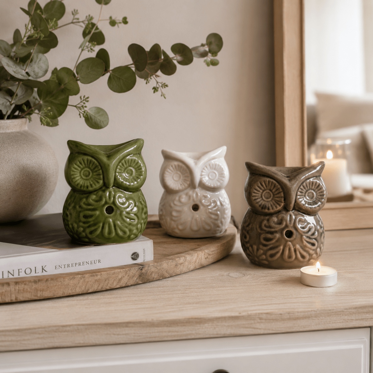 Three ceramic owl oil burners in green, cream and taupe styled on a wooden console table with eucalyptus, neutral décor and a lit tealight candle.