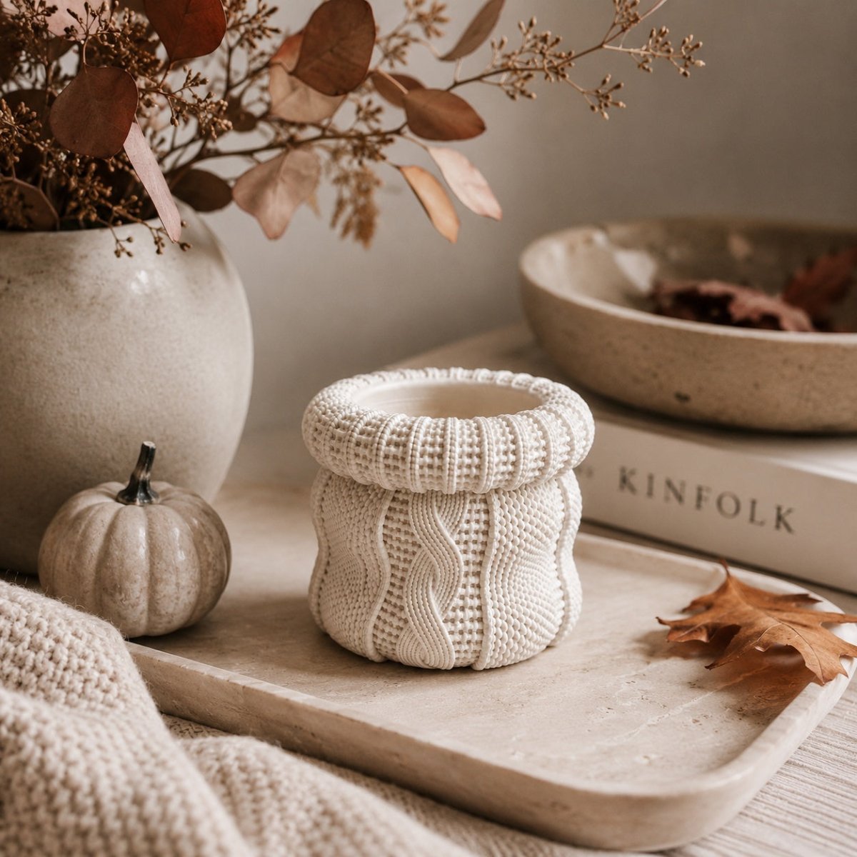 A cream-coloured stone Jar with a cable-knit pattern, styled on a wooden tray with a knit blanket, a small pumpkin, and dried botanicals.