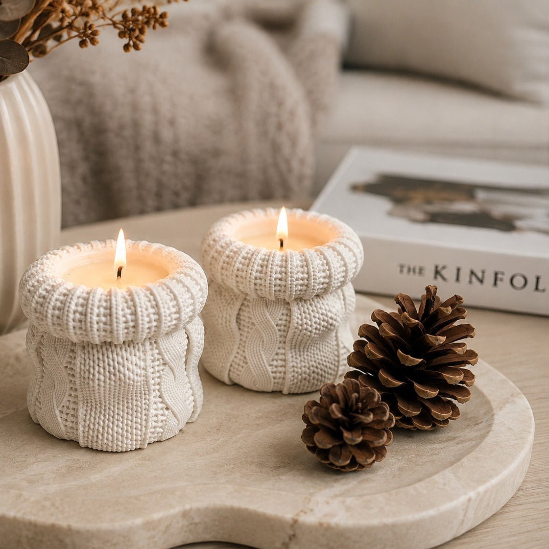 Two off white knit candles shaped like cosy sweaters, styled on a round stone tray with pinecones, a ceramic vase with dried flowers, and a book in the background. Autumn table decor