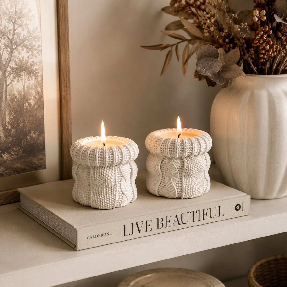 Two off white cable-knit lit candles in a handmade stone container, styled on a book on a shelf next to a ceramic vase with dried flowers. Lit Cosy Knitted Soy Candle styled for a warm autumn evening