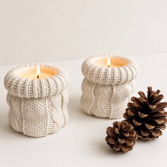 Two cream-coloured knitted candles with lit candles, styled with two pinecones on a clean white surface against a neutral off-white background.
