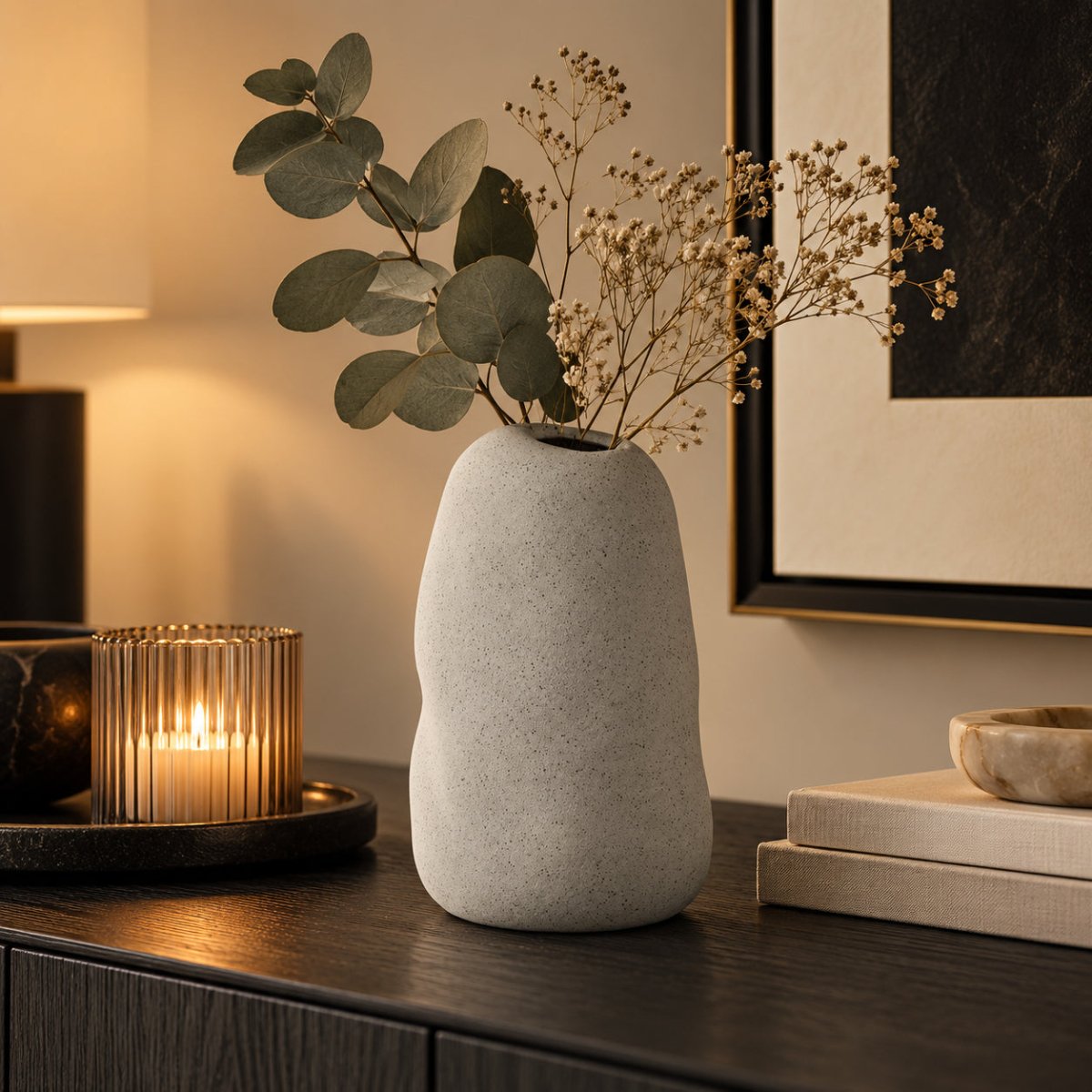 A light grey speckled ceramic vase with organic wavy contours, holding dried baby's breath and eucalyptus stems, styled on a dark wood console table in a warmly lit room.