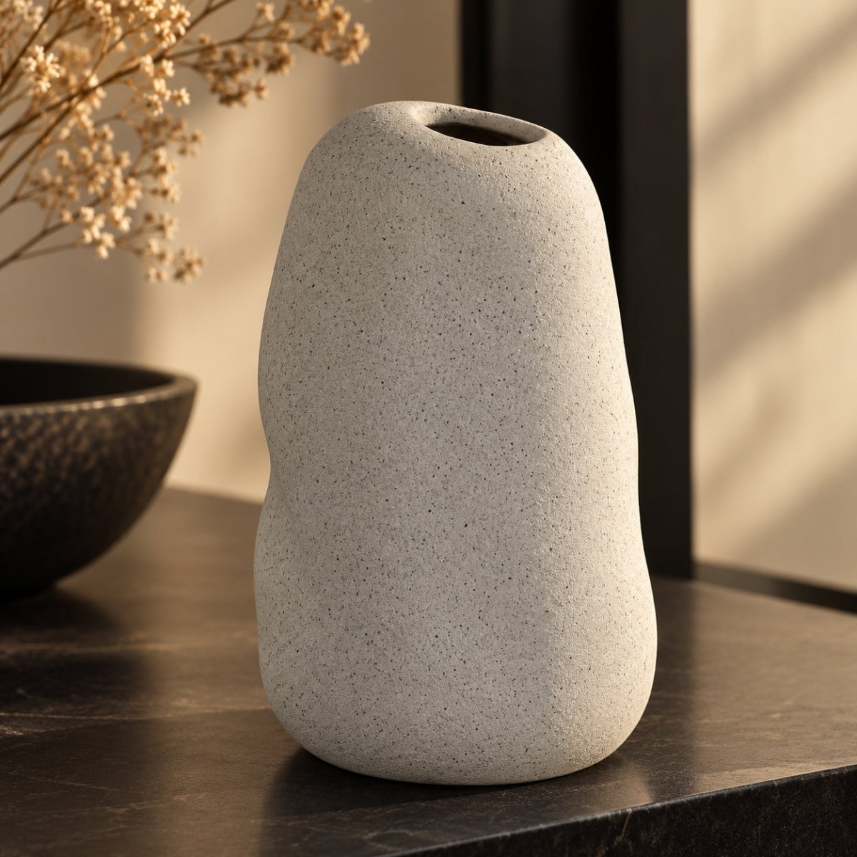 A speckled light grey ceramic vase with a rounded organic shape and a small top opening, standing on a dark tabletop in a softly lit interior setting.