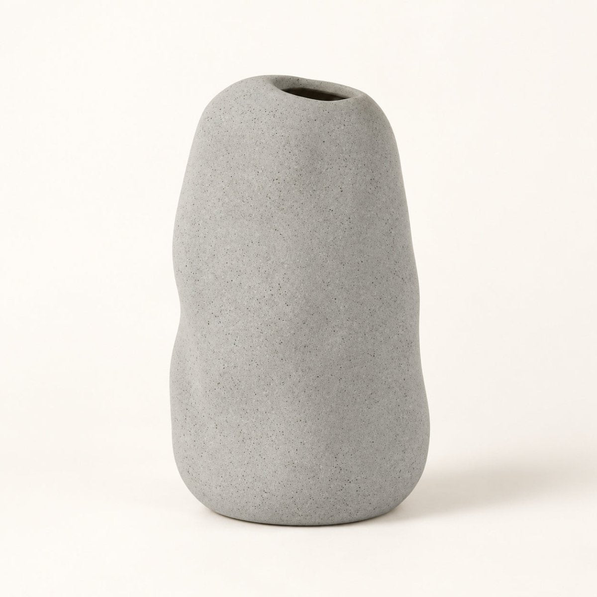 A minimalist, organic-shaped gray ceramic vase with a rough, textured matte finish and a small opening at the top, set against a clean off-white background.