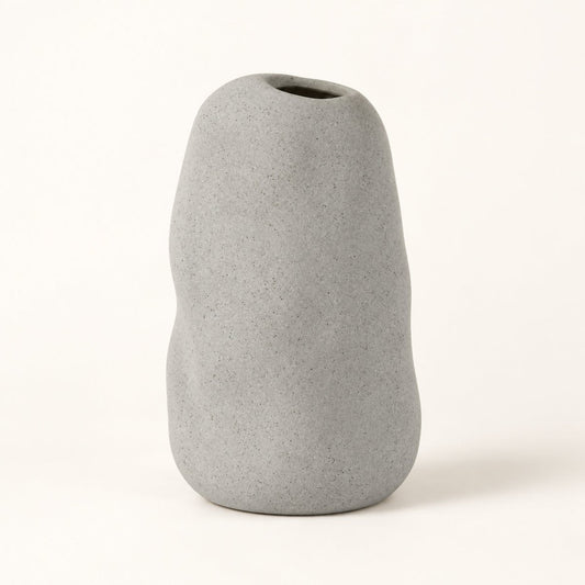 A minimalist, organic-shaped gray ceramic vase with a rough, textured matte finish and a small opening at the top, set against a clean off-white background.