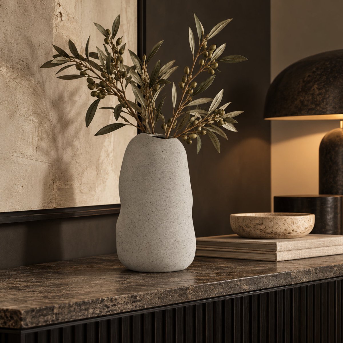 A light grey textured ceramic vase with organic wavy contours, holding olive branches, styled on a dark stone console table in a warm, modern interior setting.