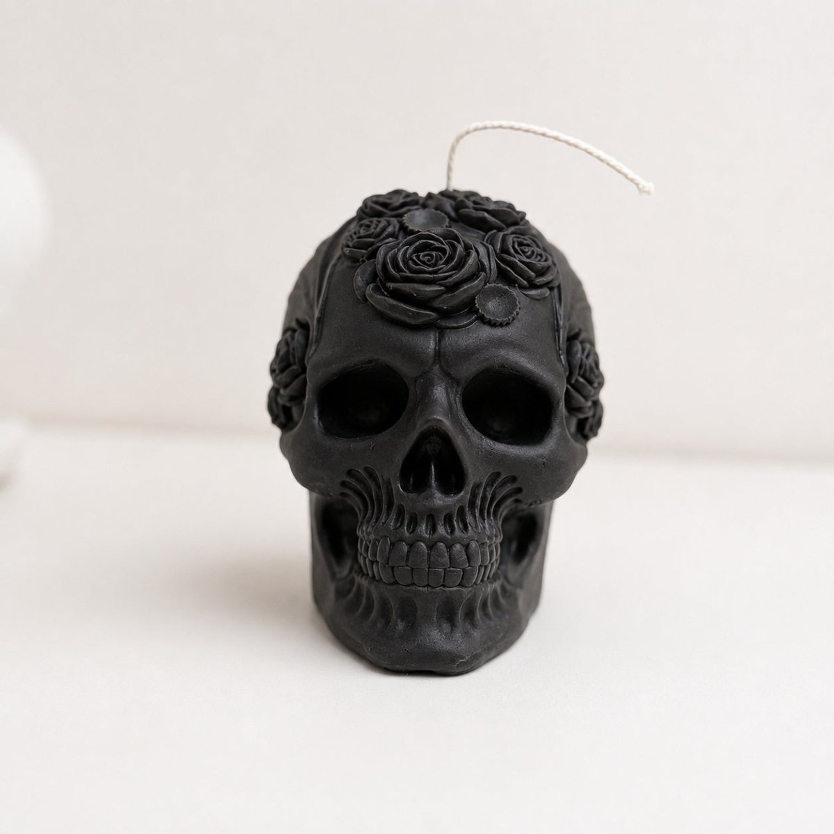 A black skull-shaped candle with rose details on top, featuring a white wick, set against a plain light background.