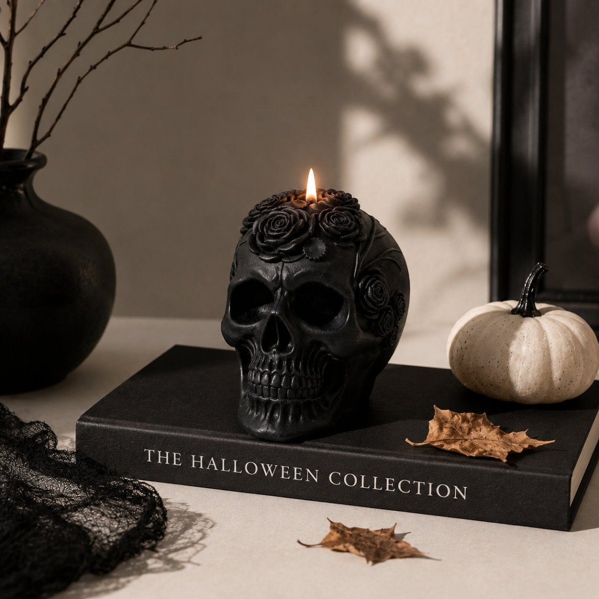A black skull-shaped candle with rose carvings on top, lit and resting on a black book labeled 'THE HALLOWEEN COLLECTION,' with a small white pumpkin, dried leaves, and black lace in a styled tabletop setting.