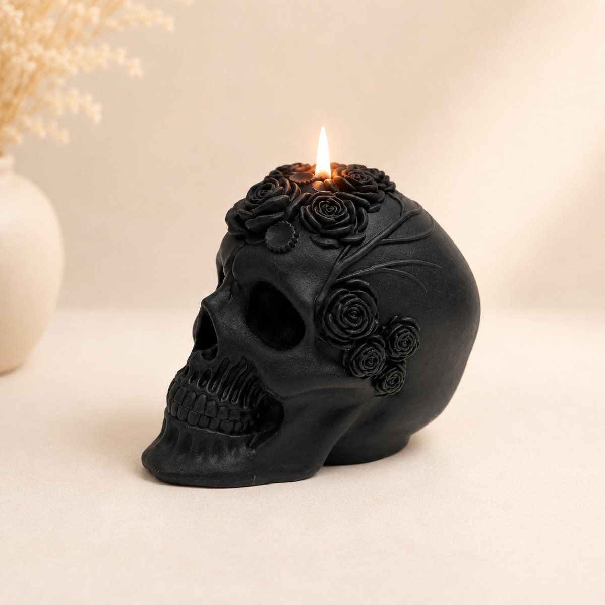 A black skull-shaped soy sculpture candle with a single flame burning from the top, decorated with raised black roses, set on a light beige surface against a soft beige background.