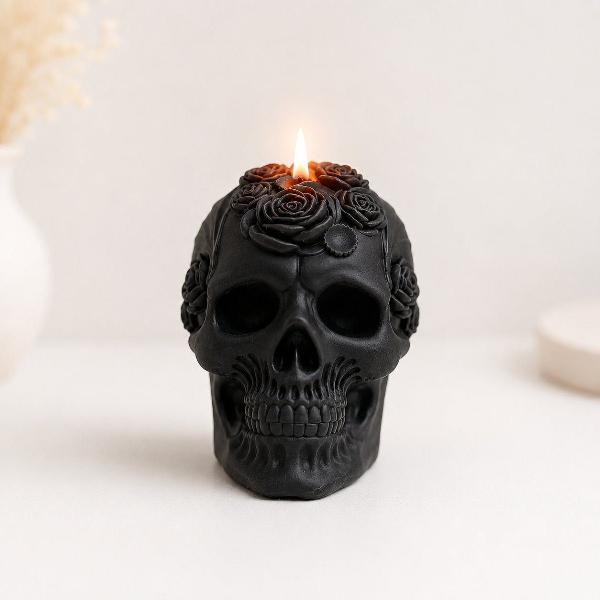 A black skull-shaped handmade candle with rose carvings on top, lit with a small flame, set against a soft white background with blurred decorative elements.