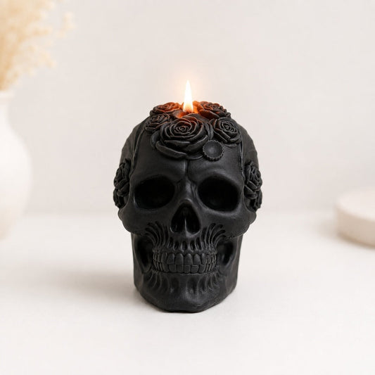 A black skull-shaped handmade candle with rose carvings on top, lit with a small flame, set against a soft white background with blurred decorative elements.