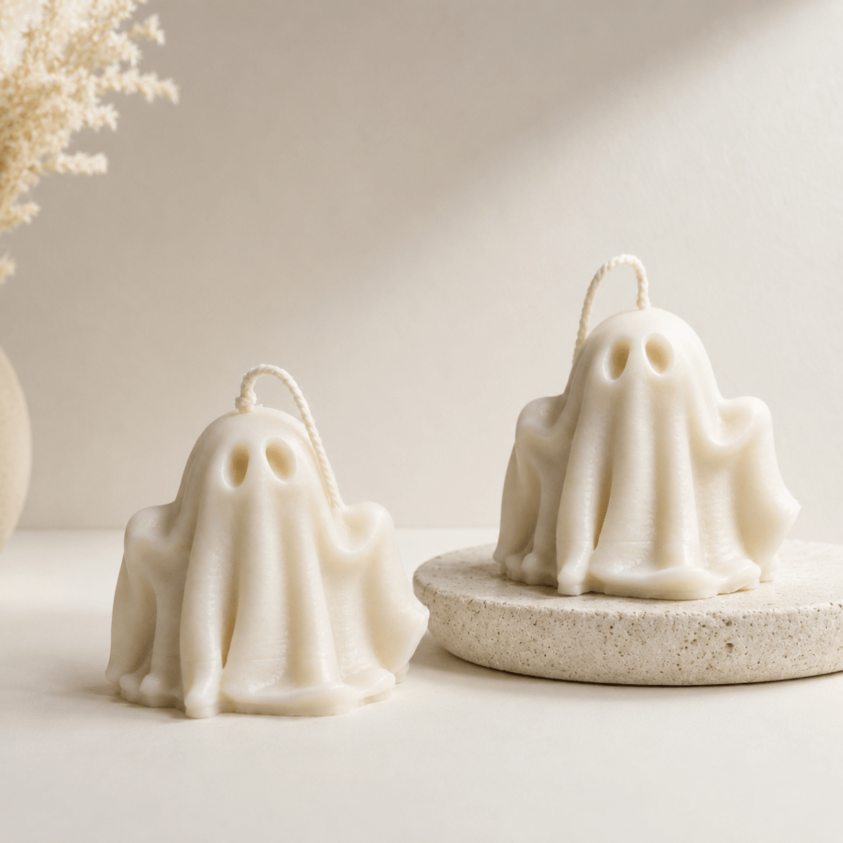 Two ghost-shaped halloween candles in an off-white cream colour, styled on a neutral beige surface with one candle resting on a round textured stone coaster. A dried floral arrangement is visible in the background on the left.