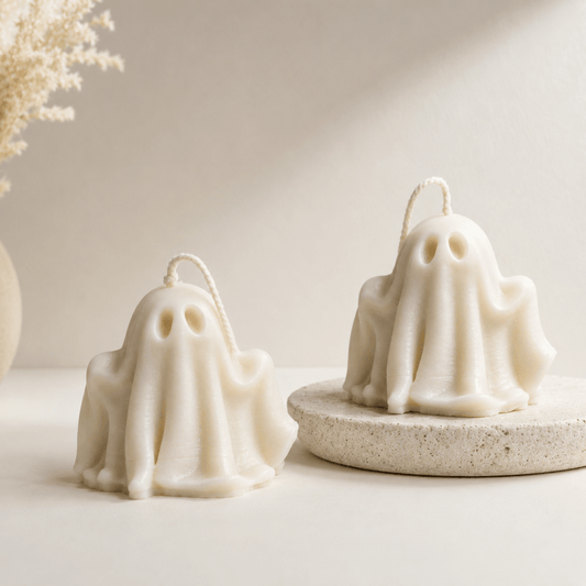 Two ghost-shaped halloween candles in an off-white cream colour, styled on a neutral beige surface with one candle resting on a round textured stone coaster. A dried floral arrangement is visible in the background on the left.