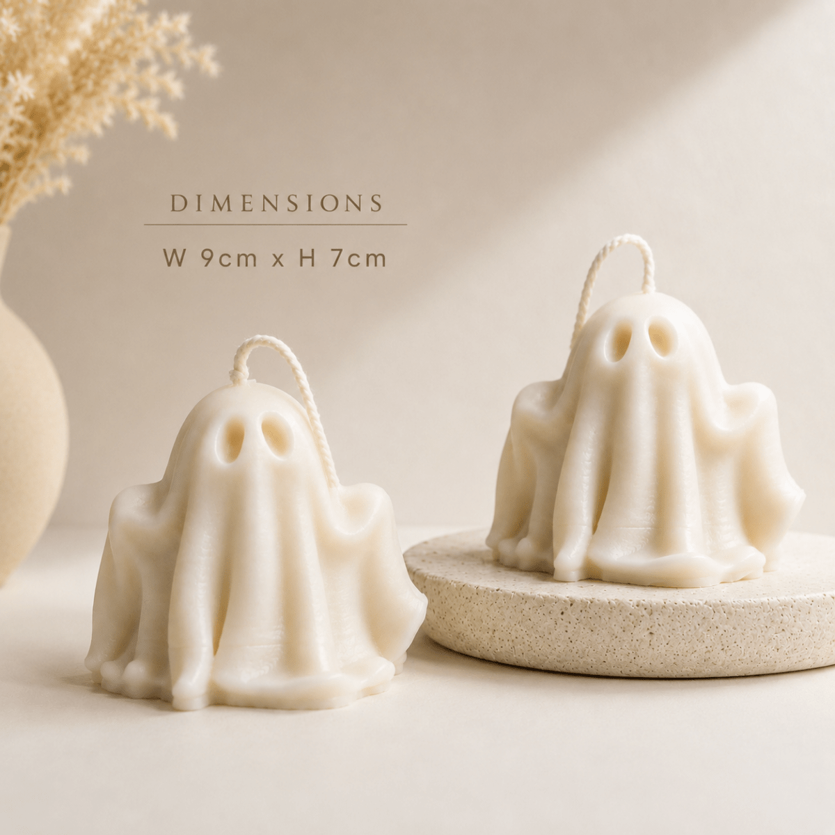 Two ghost-shaped cream candles with hanging loops, shown on a warm beige surface with dried grass decor and a stone base. Text reads “DIMENSIONS W 9cm x H 7cm.