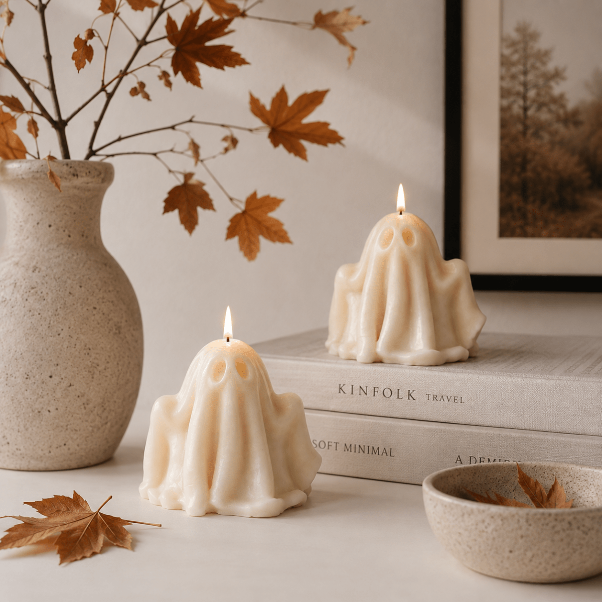 Two cream-colored ghost-shaped candles with lit flames, styled on books next to a ceramic vase with autumn leaves and a small bowl.