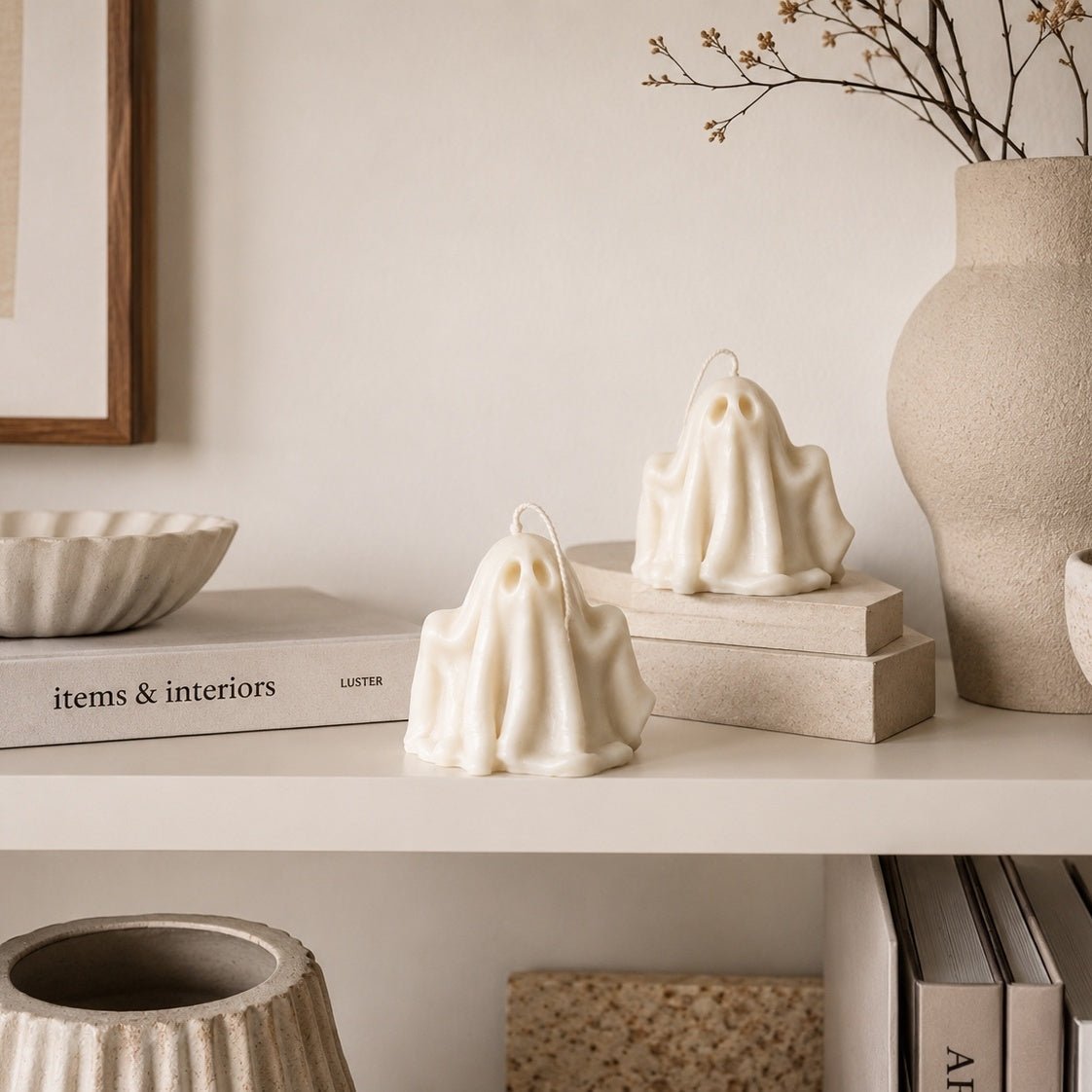 Two ghost-shaped Ivory sculpture candles styled on a shelf with neutral decor, including a ceramic vase, books, and a bowl.