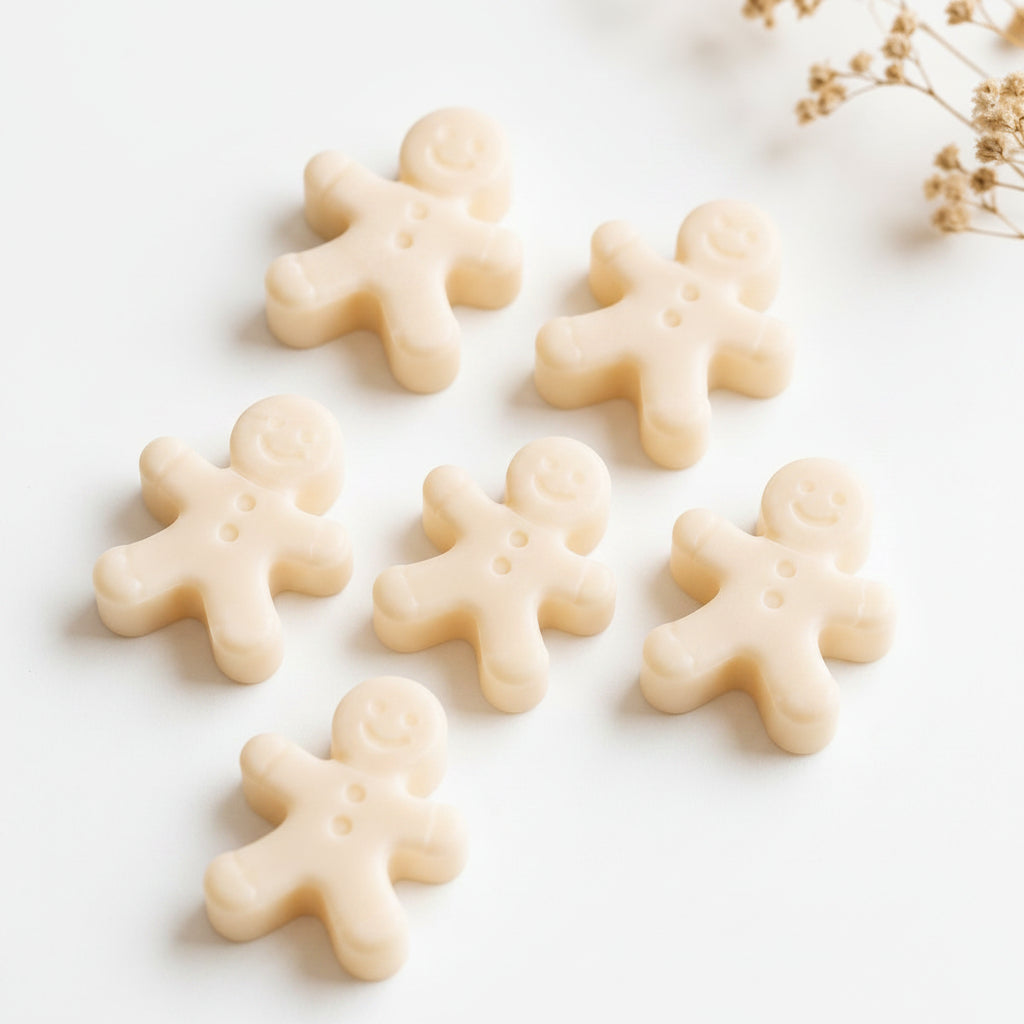 Six small gingerbread man-shaped soy wax melts in an off-white cream colour, arranged on a light surface with dried flowers in the corner.