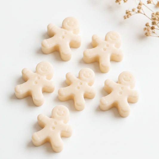 Six small gingerbread man-shaped soy wax melts in an off-white cream colour, arranged on a light surface with dried flowers in the corner.
