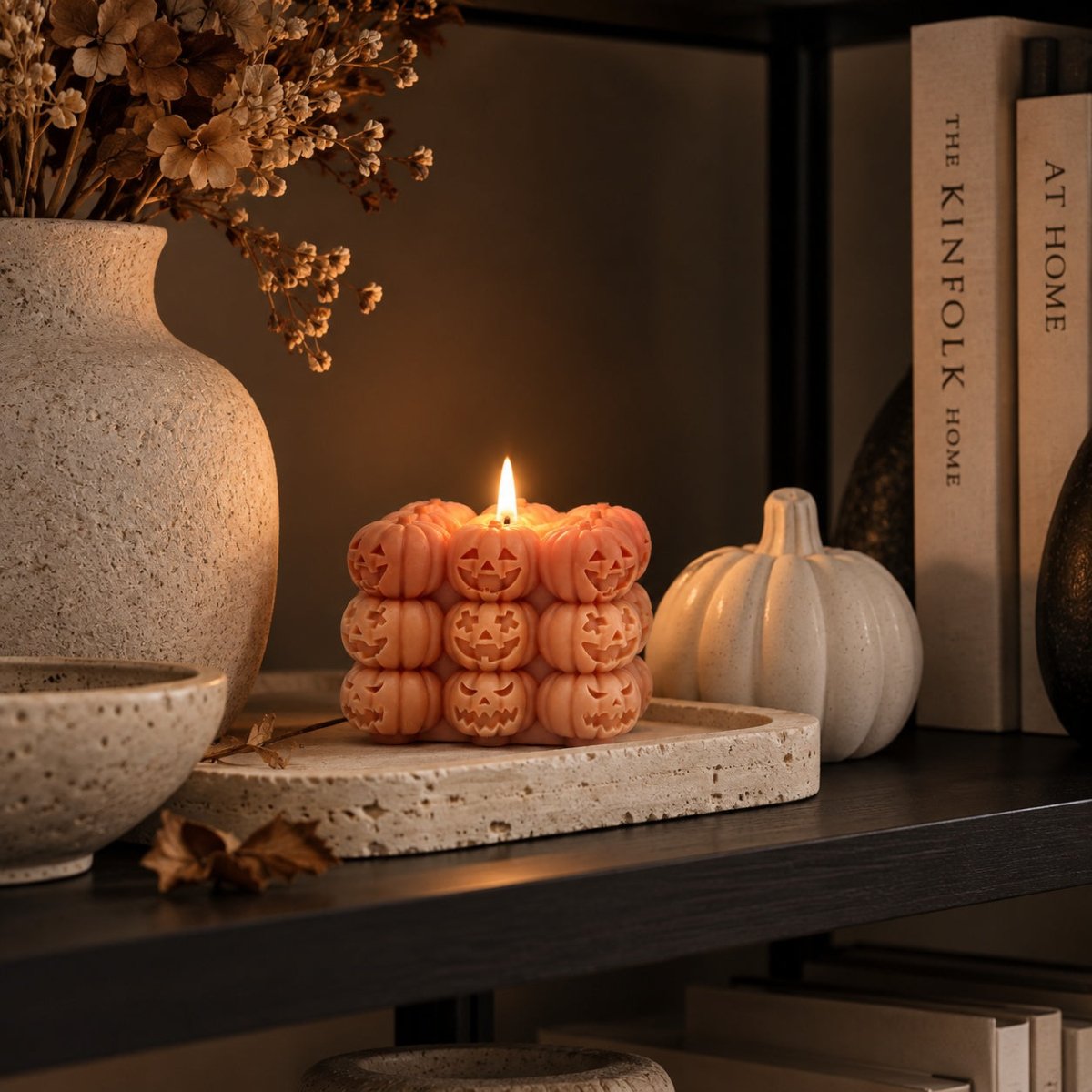 A lit candle shaped like a stack of carved jack-o'-lantern faces in a warm peachy-orange color, displayed on a stone tray on a dark shelf with dried flowers, books, and a small pumpkin in a cosy autumn indoor setting.