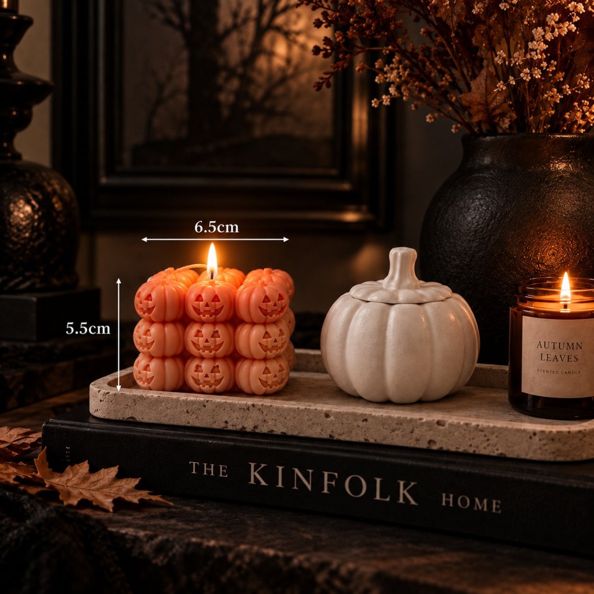 A styled tabletop scene with a lit orange jack-o'-lantern Halloween candle  stack measuring 6.5cm wide by 5.5cm tall and a white pumpkin-shaped vessel, alongside a lit jar candle labeled Autumn Leaves and decorative books and dried flowers in the background.